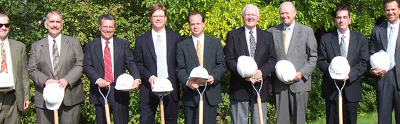 Three men in suits holding white construction helmets and shovels outdoors.